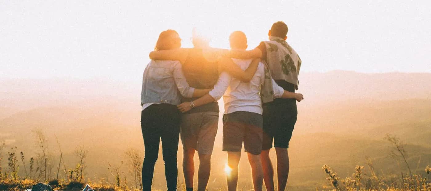 Four friends with their arms around each other, facing a golden-hour sunset over the hills